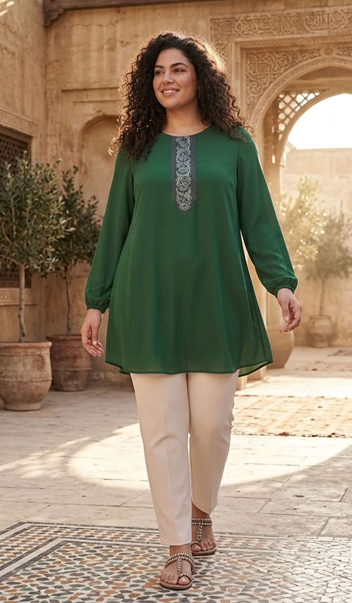 A woman modeling a green long-sleeve tunic top with a silver-gray embroidered neckline, paired with beige trousers, standing in an outdoor courtyard with stone arches.