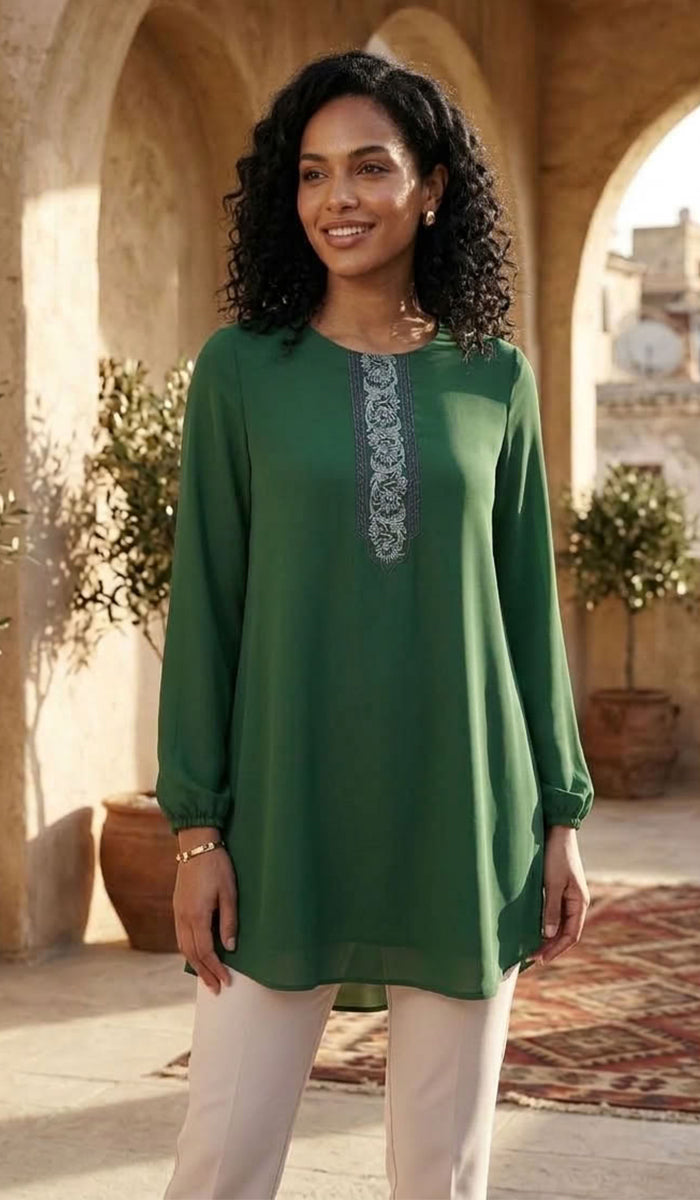A woman wearing a green long-sleeve tunic top with a decorative embroidered neckline, paired with light beige pants, standing in front of a warm stone archway background.