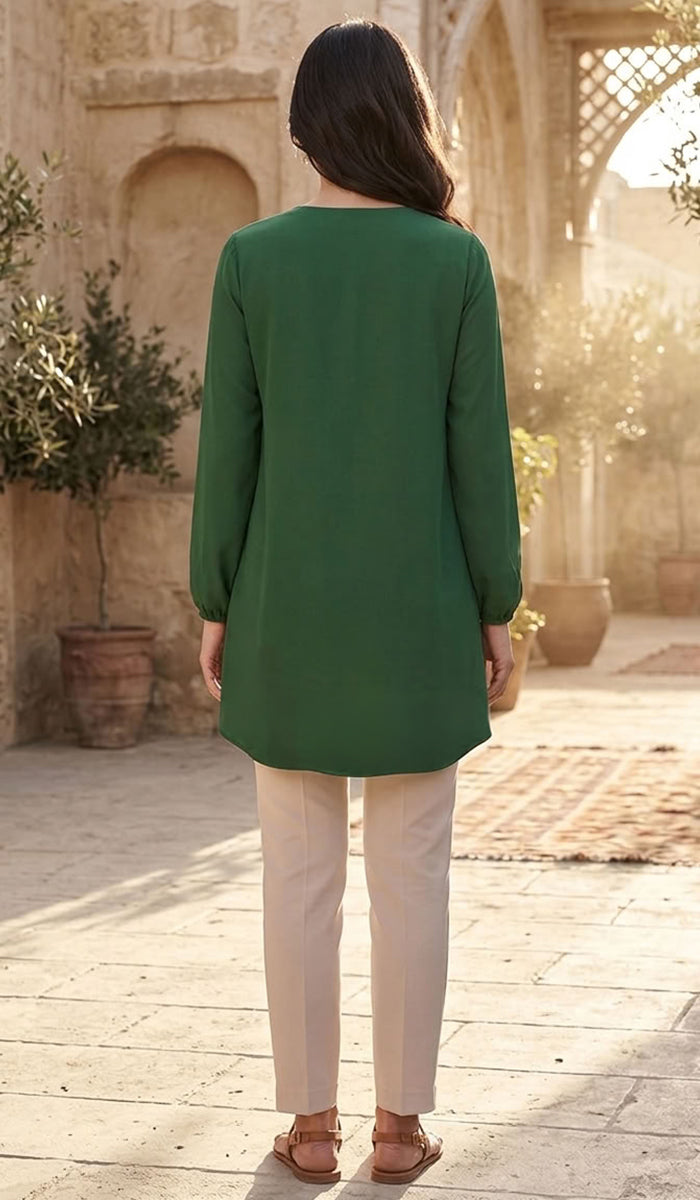 A woman viewed from behind wearing a green long-sleeve tunic top and cream pants, standing in a sunlit courtyard with stone walls and potted plants.