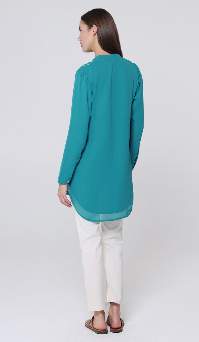 Woman wearing a teal long-sleeve top and white pants on a light gray background