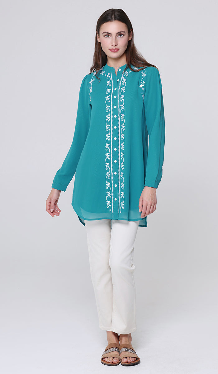 Woman wearing a teal embroidered top and white pants on a light gray background