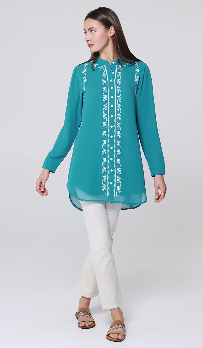 Woman wearing a teal embroidered top and white pants on a light gray background