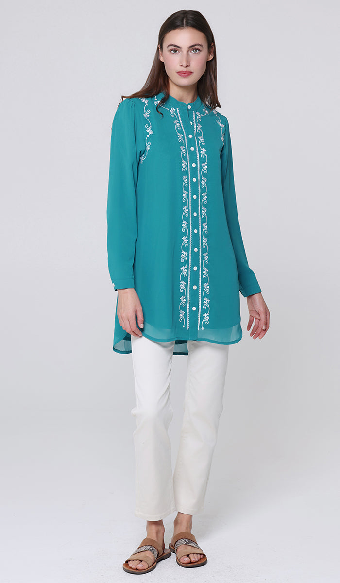 Woman wearing a teal embroidered blouse with white pants on a light gray background