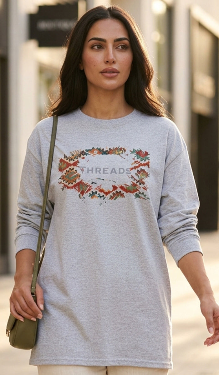 A woman wearing a light gray long-sleeve crewneck sweatshirt with a colorful floral wreath graphic and the word 'THREADS' across the chest, carrying a small olive green shoulder bag on a city street.