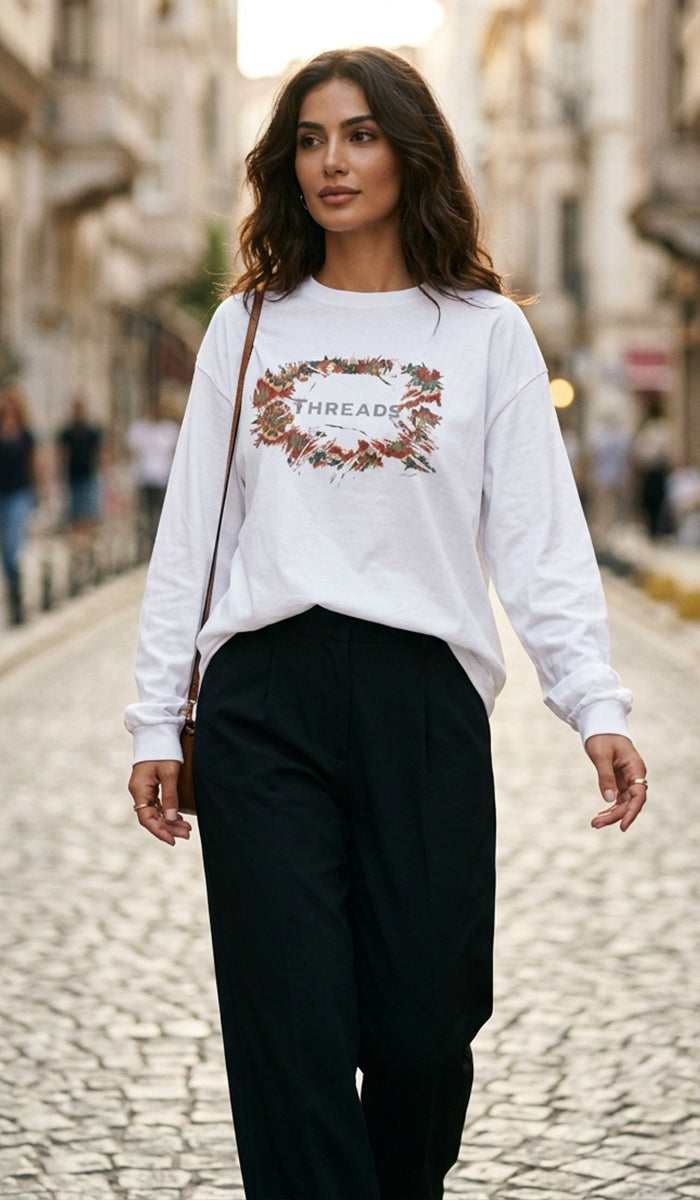 A woman walking down a cobblestone street wearing a white long-sleeve crewneck sweatshirt with a floral wreath graphic and the word 'THREAD' in the center, paired with black wide-leg pants.