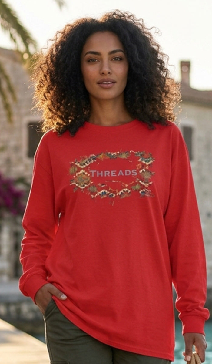 A woman with curly dark hair wearing a bright red long-sleeve crewneck sweatshirt with a 'THREADS' graphic across the chest, posed outdoors against a blurred background of buildings and palm trees.