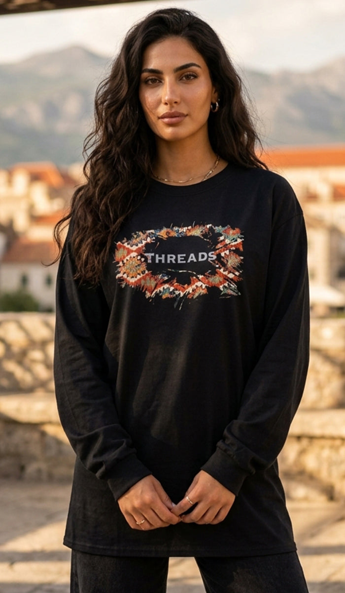 A woman wearing a black long-sleeve crewneck sweatshirt with a colorful graphic print on the chest that reads 'THREADS'. She is standing outdoors with a blurred stone wall and mountains in the background.