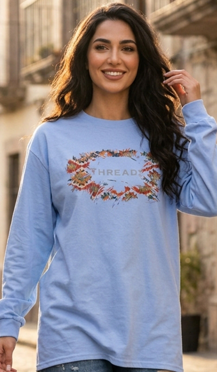 A woman wearing a light blue long-sleeve t-shirt with a colorful floral wreath graphic and the word 'THREADS' across the chest, set against a blurred outdoor background.
