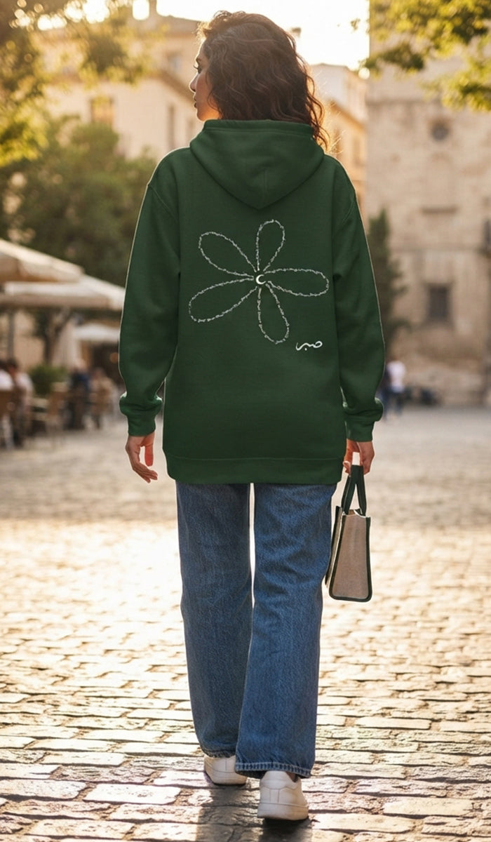 A woman viewed from behind walking on a cobblestone street, wearing a dark green hoodie with a white outlined flower graphic on the back, blue jeans, and white sneakers, while carrying a beige handbag.