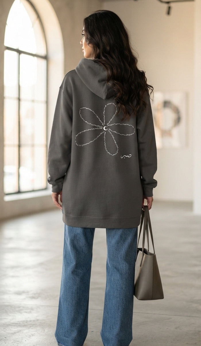 A woman stands in a bright indoor space with large arched windows, viewed from behind. She wears a dark gray oversized hoodie with a white flower outline graphic on the back, blue wide-leg jeans, and carries a taupe tote bag.