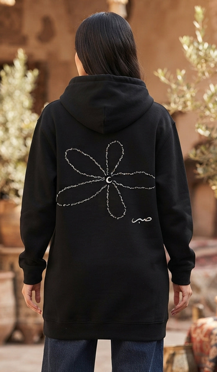A person viewed from behind wearing a black hooded sweatshirt with a large white abstract flower-like line graphic and a small white script logo on the back, paired with blue jeans in an outdoor rustic setting.