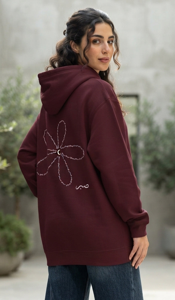 A woman wearing a burgundy hoodie with a white embroidered flower design on the back, paired with blue jeans, photographed in an outdoor setting with a blurred gray wall and greenery in the background.