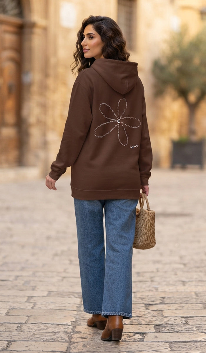 A woman wearing a brown hoodie with a white flower graphic on the back, paired with blue jeans and brown boots, standing on a cobblestone street.