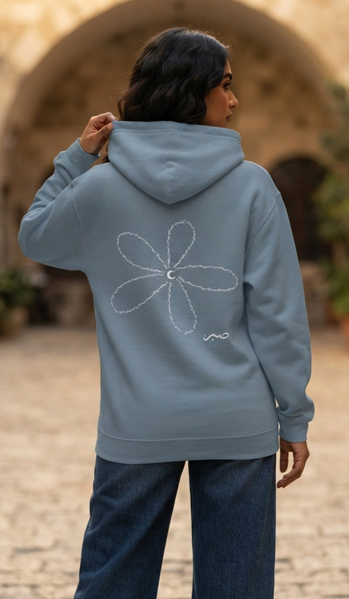 A person wearing a dusty blue hoodie with a white hand-drawn flower graphic on the back, standing outdoors in front of a blurred stone archway.