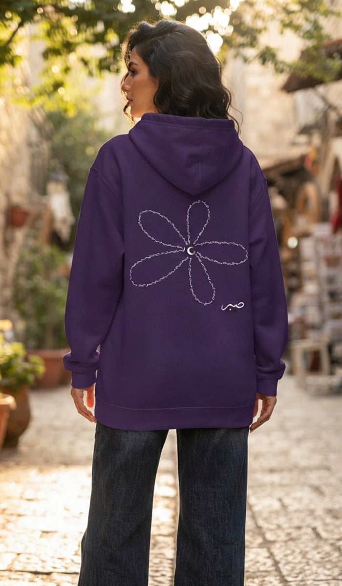 A woman viewed from behind wearing a purple hoodie with a white outlined flower graphic on the back, paired with dark jeans, standing outdoors on a sunlit cobblestone path.