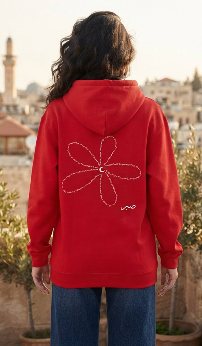 A person viewed from behind wearing a bright red hoodie with a large white flower outline and a small white script detail on the back, set against an outdoor cityscape background.
