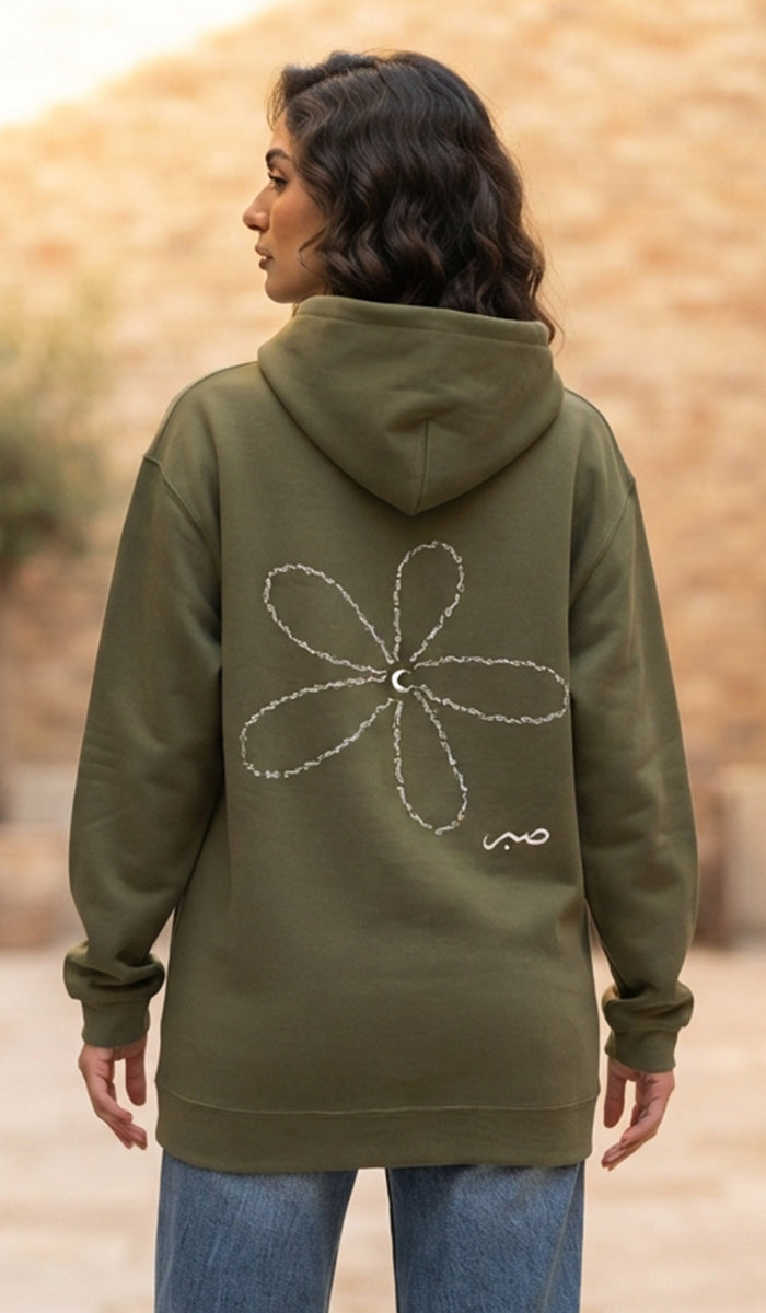 A woman viewed from behind wearing an olive green hooded sweatshirt with a large white flower graphic and a small white script logo on the back, paired with blue jeans against a blurred outdoor background.