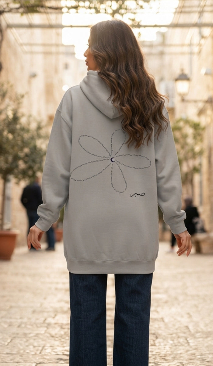 A woman viewed from behind wearing a light gray oversized hoodie with a large outlined flower graphic and a small script detail on the back, paired with dark blue jeans in an outdoor street setting.
