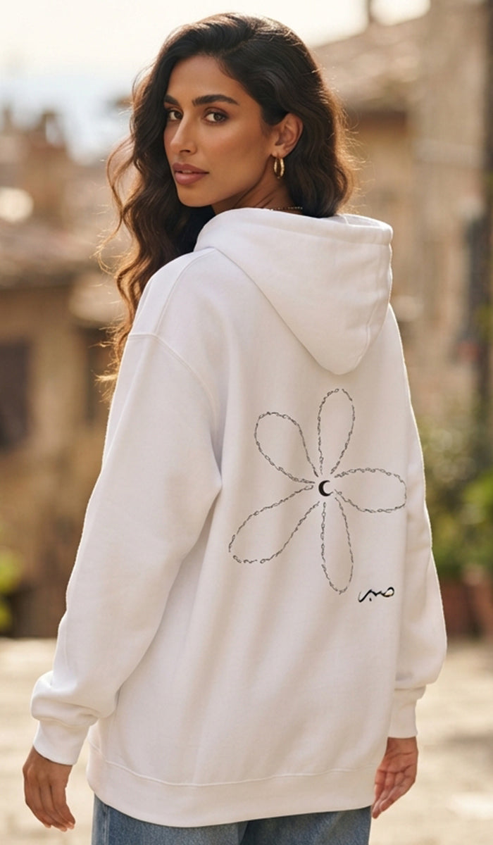 A woman wearing a white pullover hoodie with a black line-art flower graphic and a small signature-style logo on the back, photographed outdoors against a softly blurred stone building background.