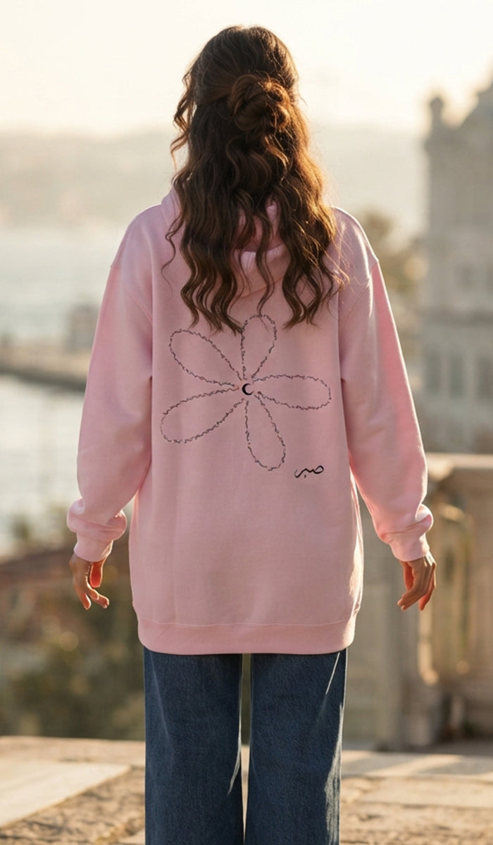 A woman viewed from behind wearing a light pink hoodie with a large outlined flower graphic on the back, paired with dark blue jeans, standing outdoors against a softly blurred background.