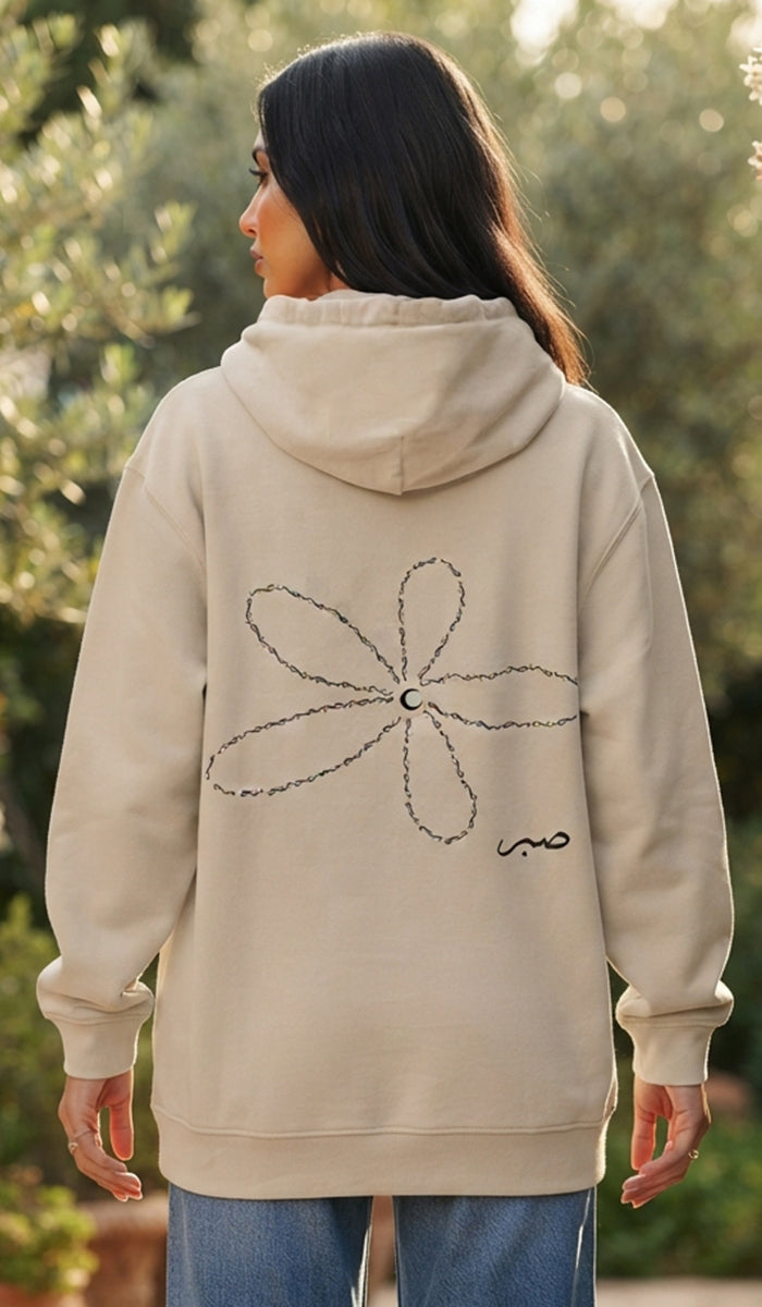 A woman viewed from behind wearing a beige hoodie with a large flower graphic and a small script logo on the back, paired with blue jeans in an outdoor setting.