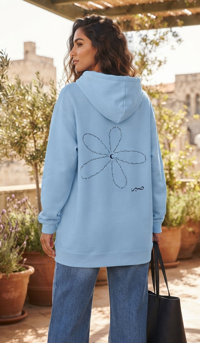 A woman wearing a light blue hoodie with a black line-art flower graphic on the back, standing on a sunlit outdoor terrace with potted plants and buildings in the background.