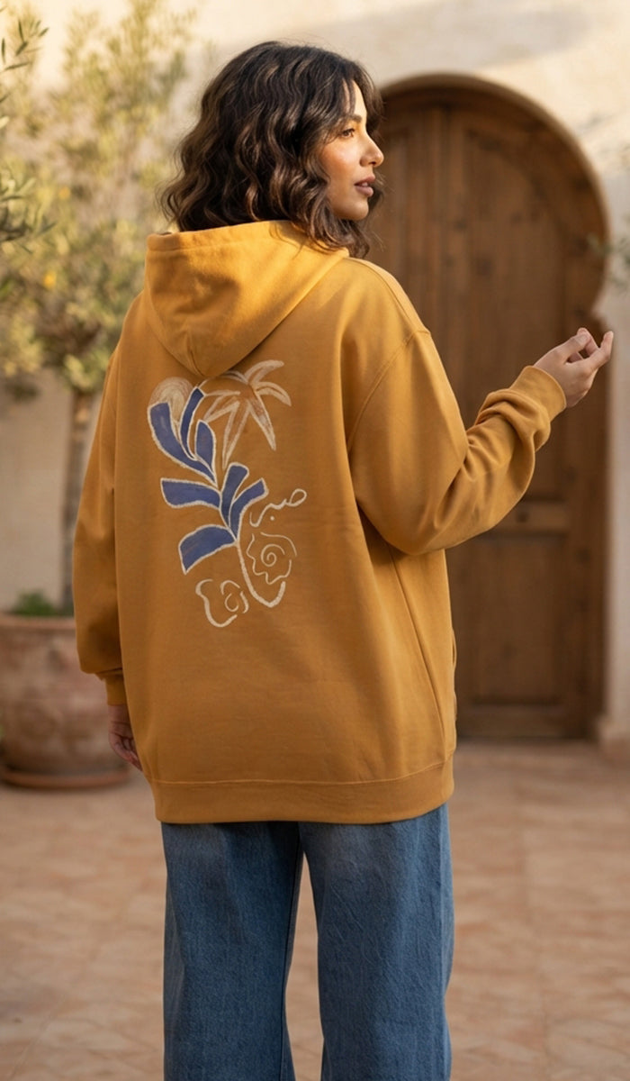A woman wearing an oversized mustard yellow hoodie with a blue and white floral graphic on the back, paired with blue jeans, standing in front of a rustic wooden door.