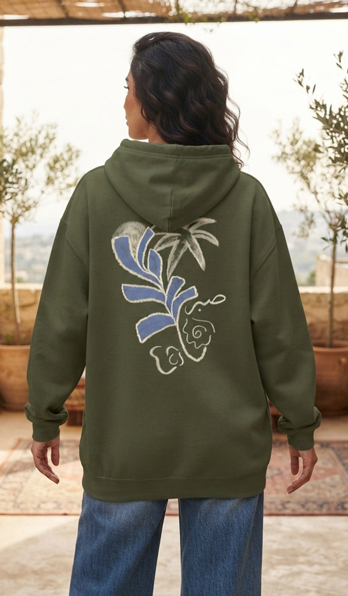 A woman viewed from behind wearing an olive green hoodie with a large blue and white floral graphic on the back, paired with blue jeans in an outdoor setting.
