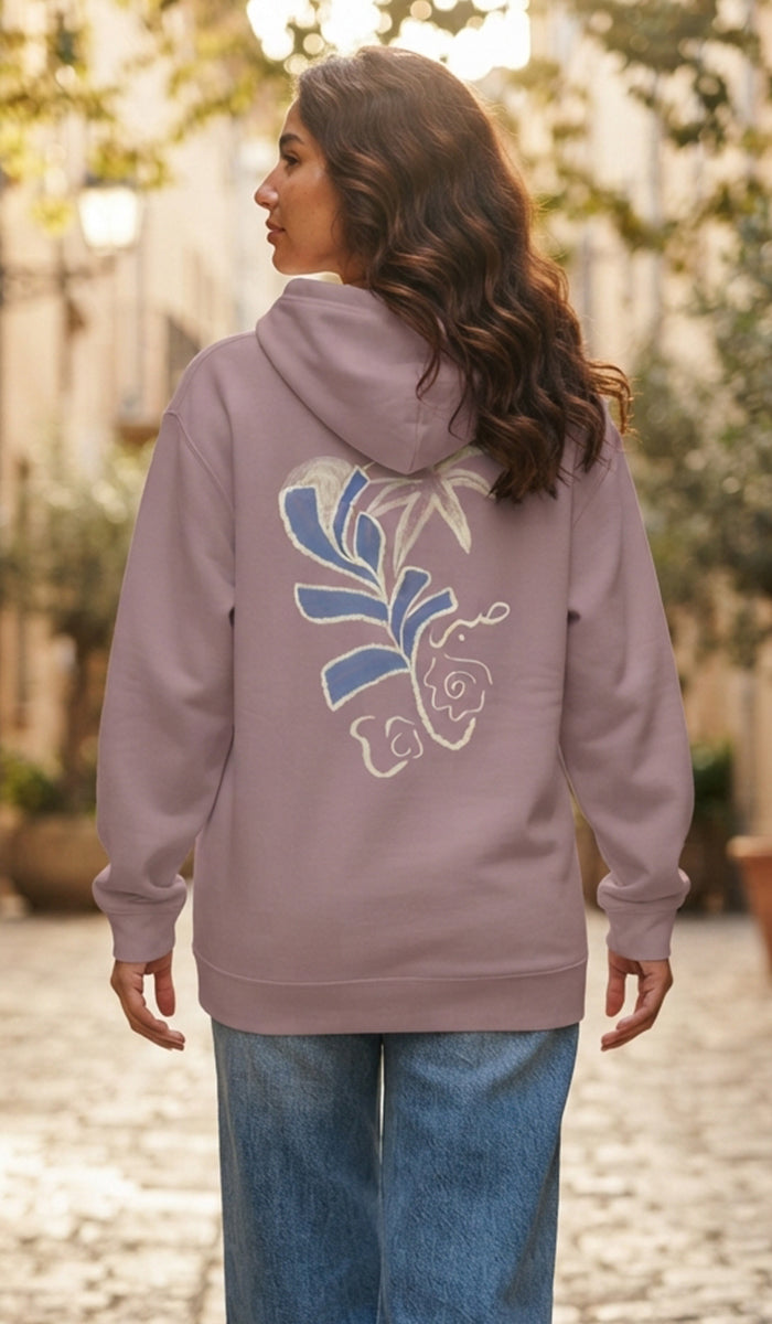A woman wearing a lavender hoodie with a blue and white floral graphic on the back, paired with blue jeans, standing in an outdoor setting with blurred trees and buildings in the background.