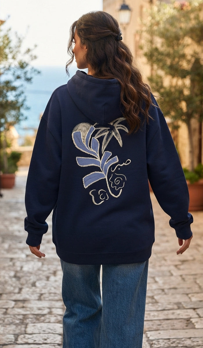 A woman viewed from behind wearing a navy blue hoodie with a large blue and white floral graphic on the back, paired with blue jeans, walking on a cobblestone street.