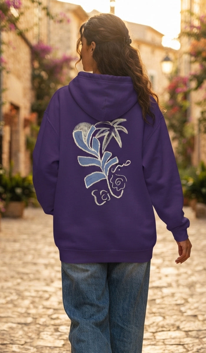 A woman viewed from behind wearing a purple hoodie with a large blue and white floral graphic on the back, paired with blue jeans, walking on a cobblestone street.