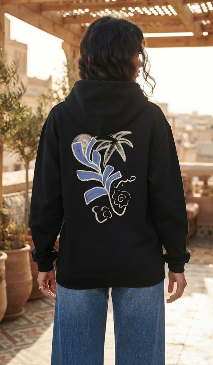 A woman viewed from behind wearing a black hoodie with a large blue, white, and silver floral and palm tree graphic on the back, paired with blue jeans, set against an outdoor patio background.