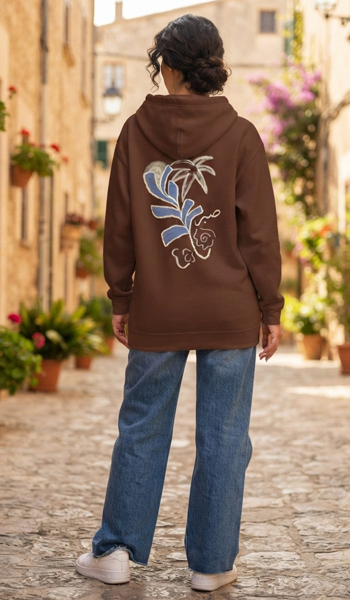 A woman viewed from behind wearing a brown hoodie with a large blue and white floral graphic on the back, paired with blue jeans and white sneakers, standing on a cobblestone street in a sunny village setting.