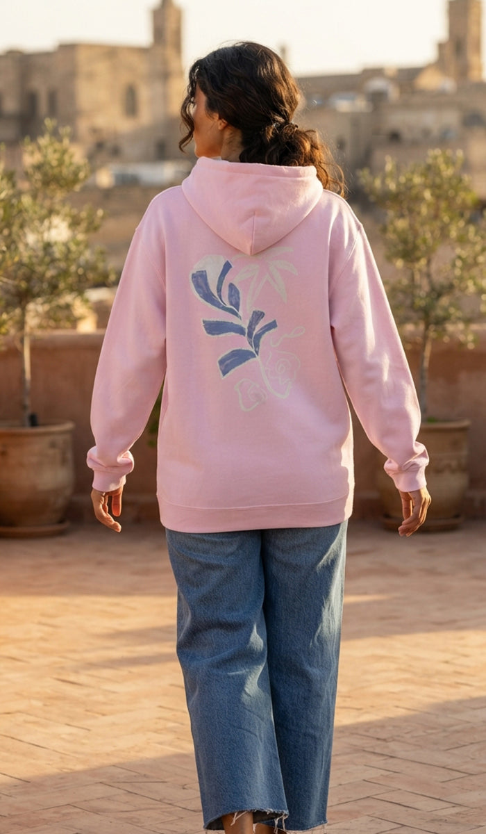 A woman viewed from behind wearing a light pink hoodie with a blue floral graphic on the back, paired with wide-leg blue jeans, standing on a sunlit outdoor terrace with warm-toned buildings in the background.