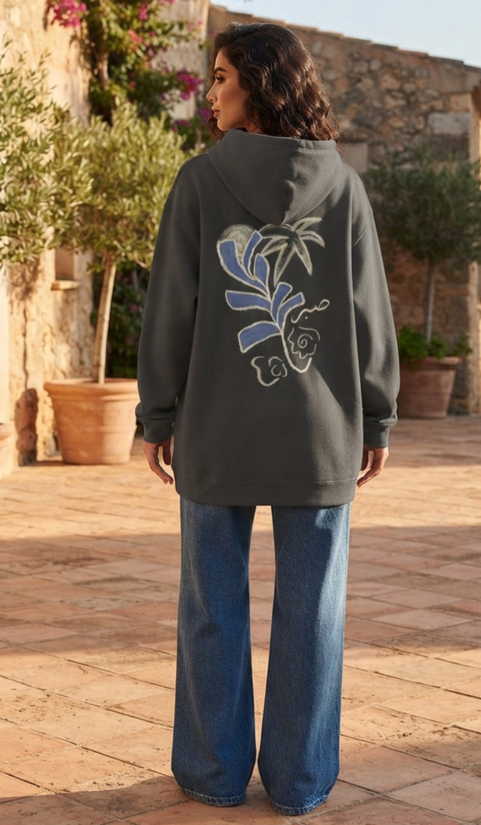 A woman stands outdoors on a stone patio wearing a dark gray oversized hoodie with a large blue and white floral graphic on the back, paired with wide-leg blue jeans.