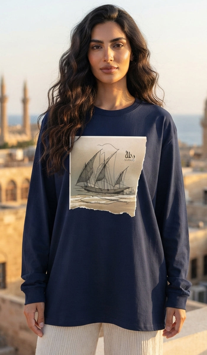 A woman wearing a navy blue long-sleeve t-shirt with a vintage-style sailboat graphic on the front, set against a warm, sunlit background of historic buildings and a sea view.