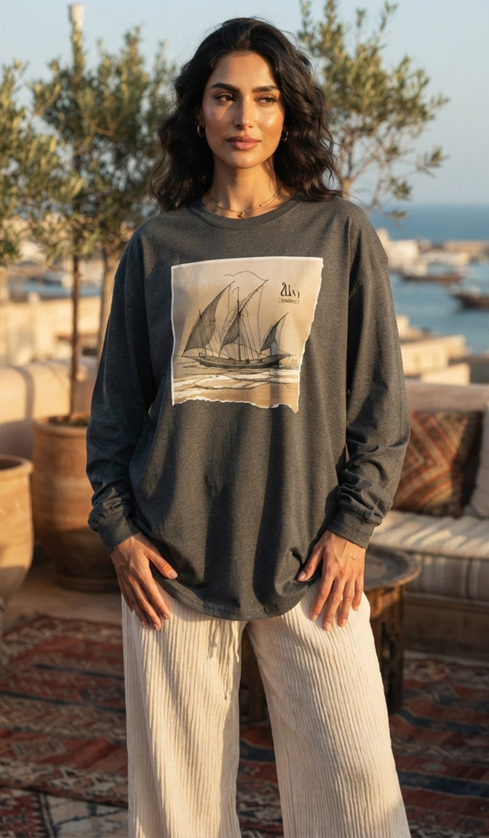 A woman stands on a sunlit terrace wearing a dark charcoal long-sleeve top with a large vintage sailboat graphic on the front, paired with light beige wide-leg pants.