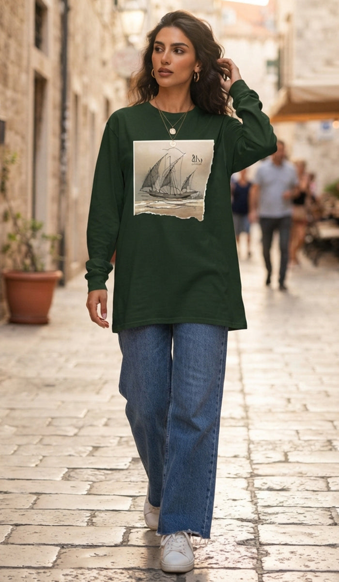 A woman walking down a cobblestone street wearing a dark green long-sleeve t-shirt with a vintage sailboat graphic on the front, paired with wide-leg blue jeans and white sneakers.