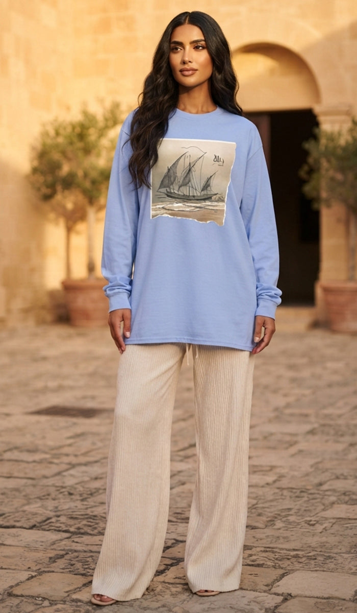 A woman stands outdoors wearing a light blue long-sleeve top with a large rectangular sailboat graphic on the front, paired with wide-leg cream pants.