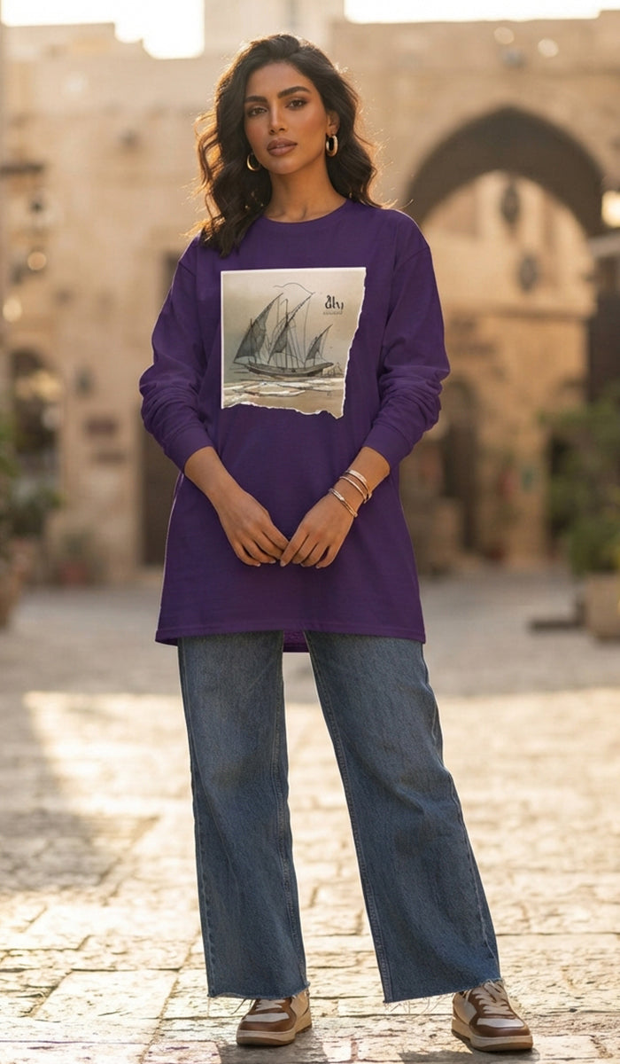 A woman wearing a purple long-sleeve tunic top with a vintage sailboat graphic print on the front, paired with wide-leg blue jeans and brown shoes, standing in an outdoor stone alleyway.