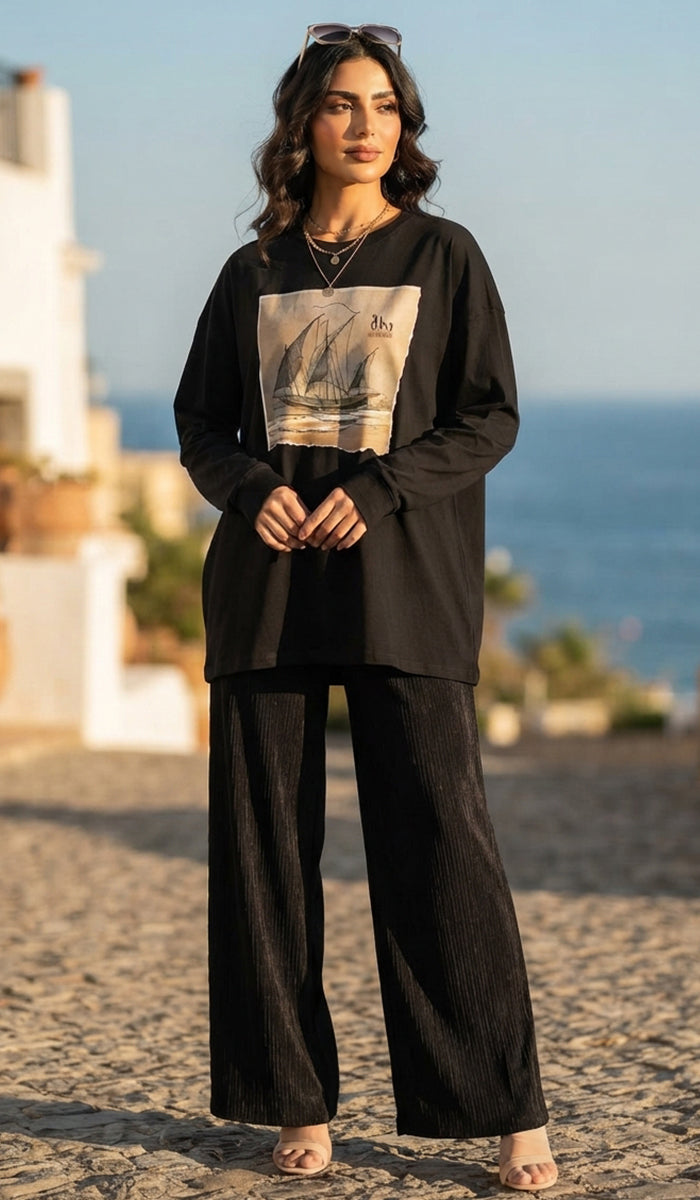 A woman stands outdoors on a sunlit stone path near the sea, wearing a loose black long-sleeve top with a large rectangular sailboat graphic on the front and matching black wide-leg pants.