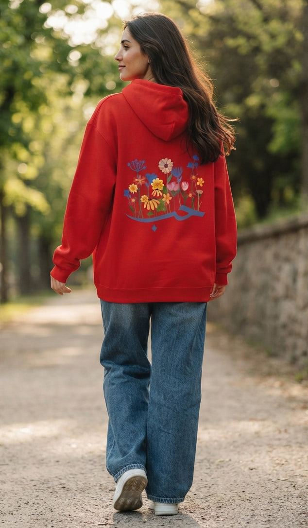A woman wearing a bright red hoodie with a colorful floral embroidery on the back, paired with blue jeans, walking on an outdoor path.