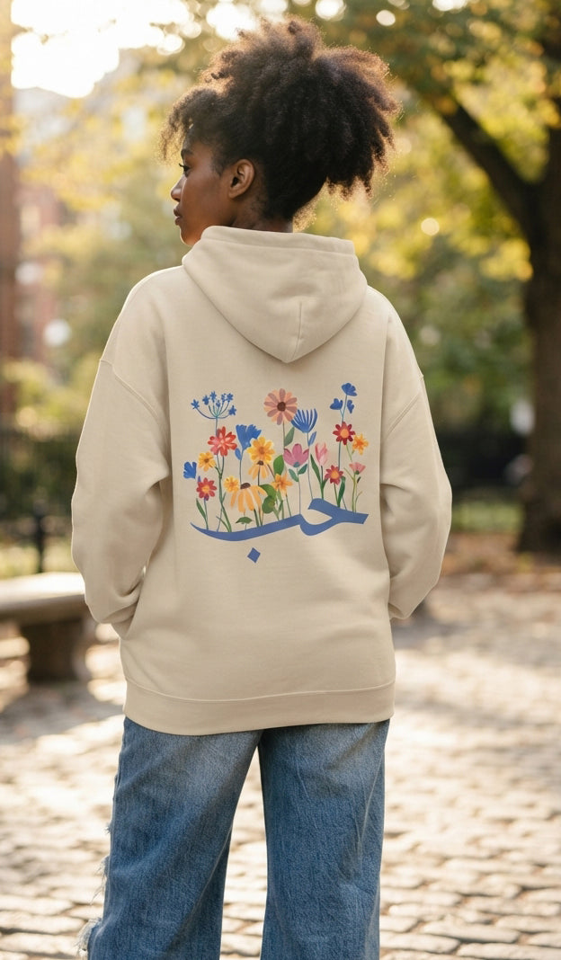 A person standing outdoors in a park wearing a cream-colored hoodie with a colorful floral and Arabic calligraphy graphic on the back, paired with blue jeans.