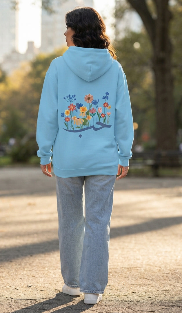 A woman viewed from behind wearing a light blue hoodie with a colorful floral and bird graphic on the back, paired with light-wash jeans and white sneakers, standing outdoors on a paved path.