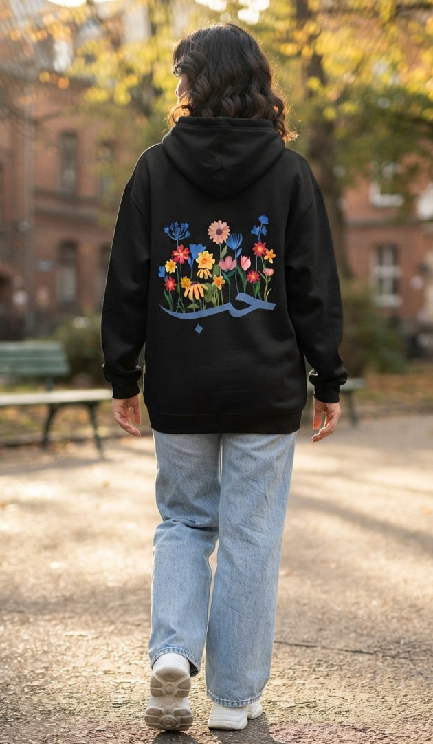 A person viewed from behind wearing a black hoodie with a colorful floral graphic on the back, paired with light blue jeans and white sneakers, walking outdoors on a path with trees and buildings in the background.