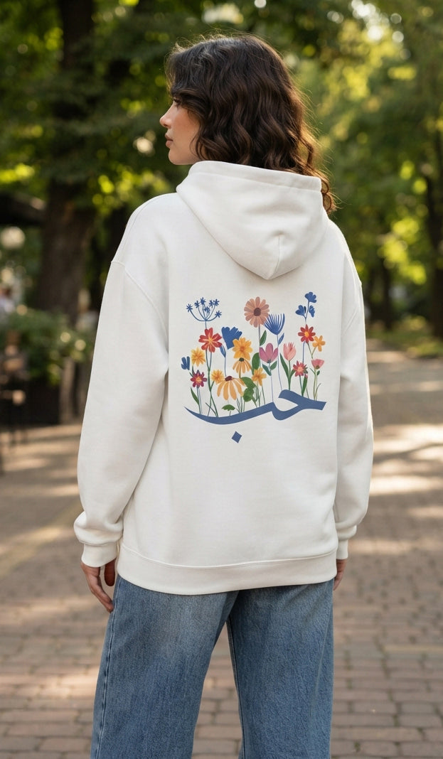 A woman viewed from behind wearing an off-white hoodie with a colorful floral and Arabic calligraphy graphic on the back, paired with blue jeans, standing outdoors on a paved path with trees in the background.