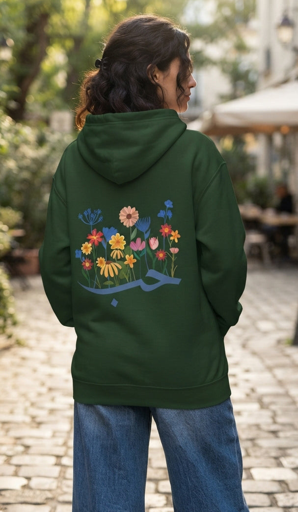 A woman viewed from behind wearing a dark green hoodie with a colorful floral and Arabic calligraphy graphic on the back, paired with blue jeans, standing on a cobblestone path.