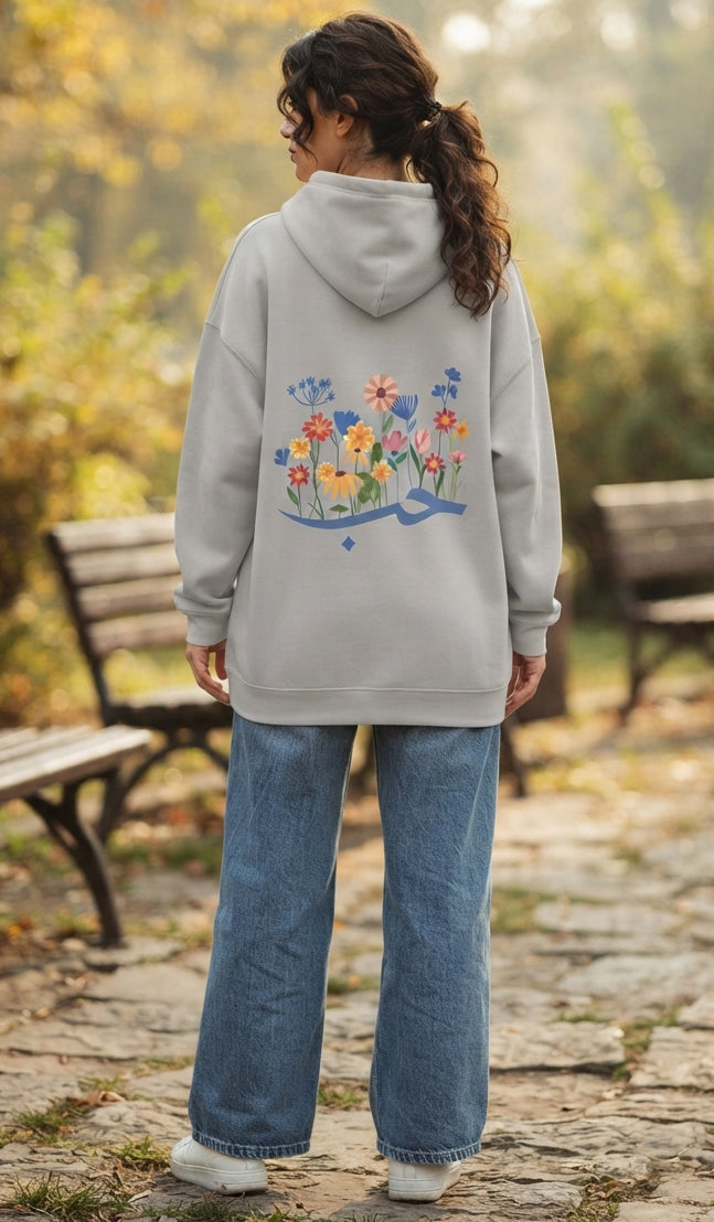 A woman viewed from behind wearing a light gray hoodie with a colorful floral graphic on the back, paired with wide-leg blue jeans and white sneakers, standing on a stone path in an outdoor park setting.