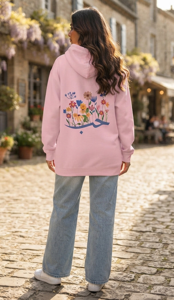 A woman viewed from behind wearing a light pink hoodie with a colorful floral and butterfly graphic on the back, paired with light blue jeans and white sneakers, standing on a cobblestone street.