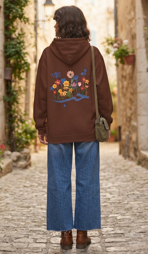 A woman viewed from behind wearing a brown hoodie with a colorful floral and bird graphic on the back, paired with wide-leg blue jeans and brown boots, standing on a cobblestone street.